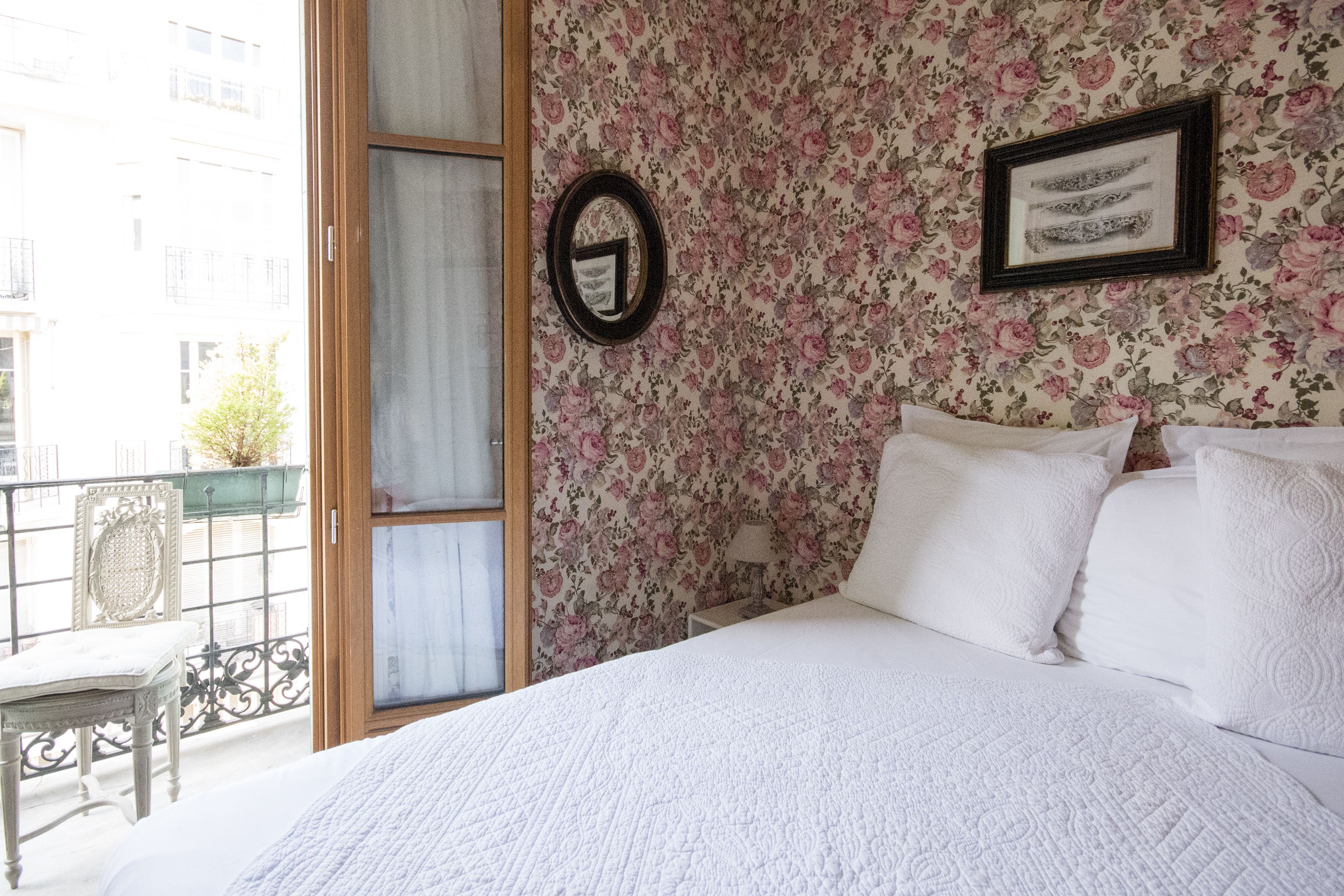 A bedroom with a bed, floral wallpaper, a framed picture, and a window with a view of the outside.