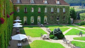 A building with a green ivy-covered exterior, a courtyard with tables and chairs, and a large tree in the center. - Les Ursulines (Autun)