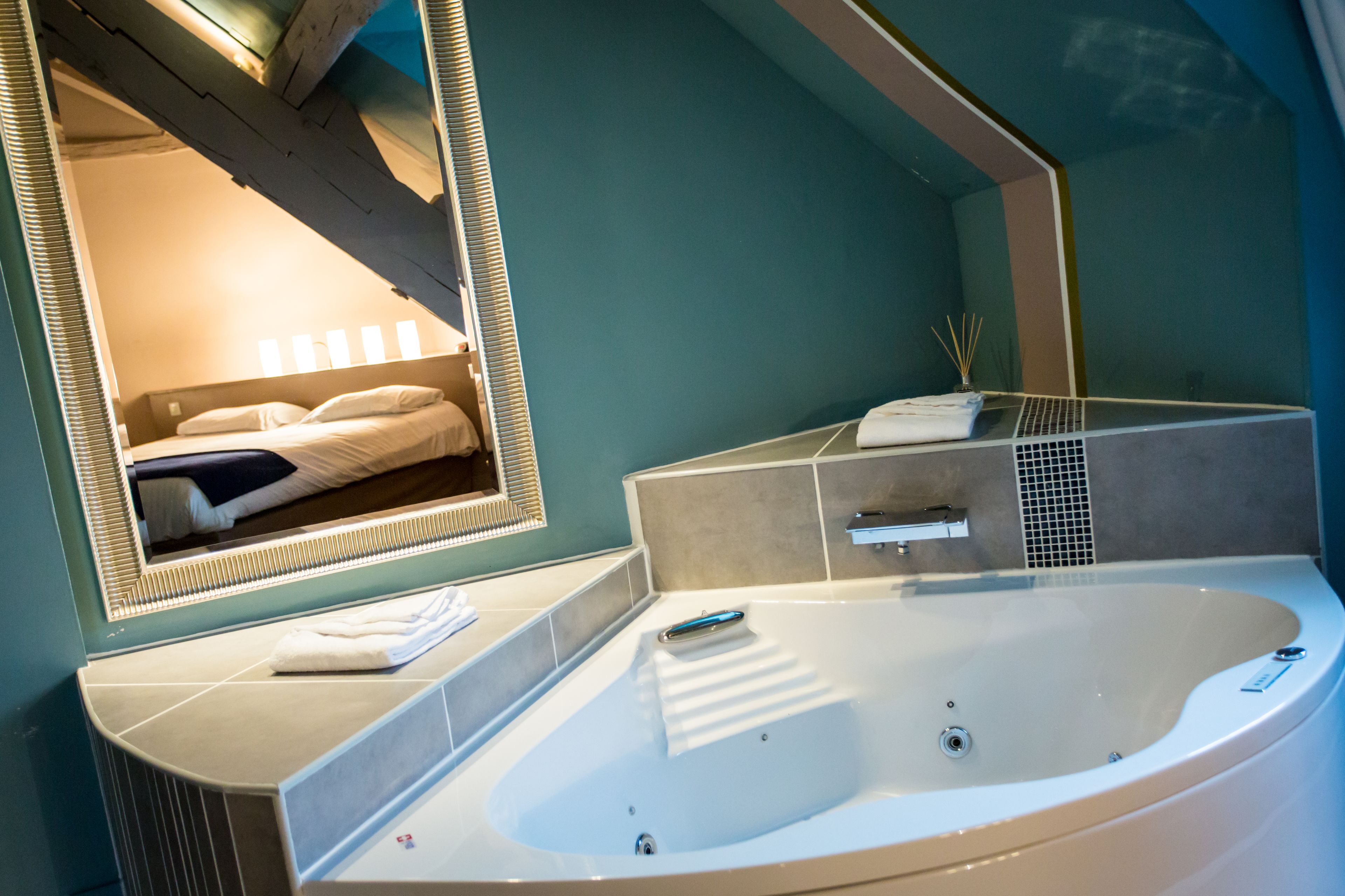 A modern bathroom with a large bathtub, a mirror, and a bed visible through the mirror.