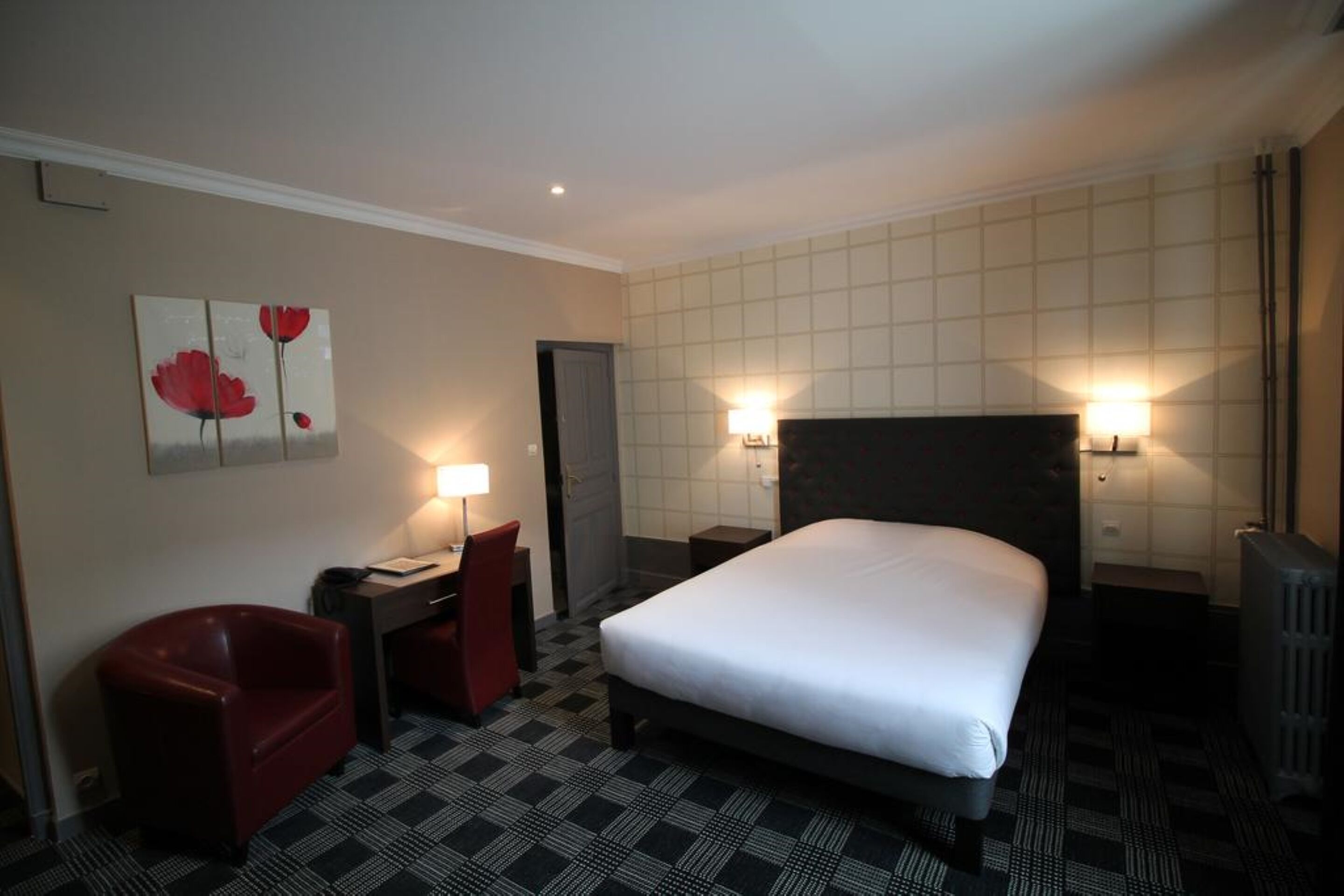 A hotel room with a bed, a desk with a chair, a lamp, and a painting on the wall.