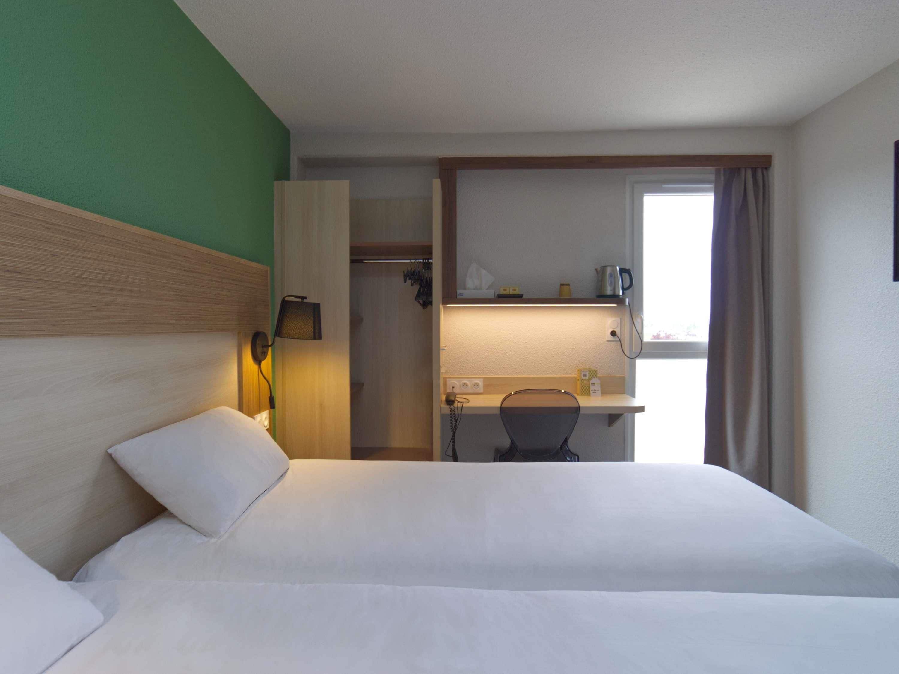 Une chambre d’hôtel avec un lit, un bureau et une chaise. Il y a une fenêtre avec des rideaux, une étagère avec des objets et une porte donnant sur une salle de bain.