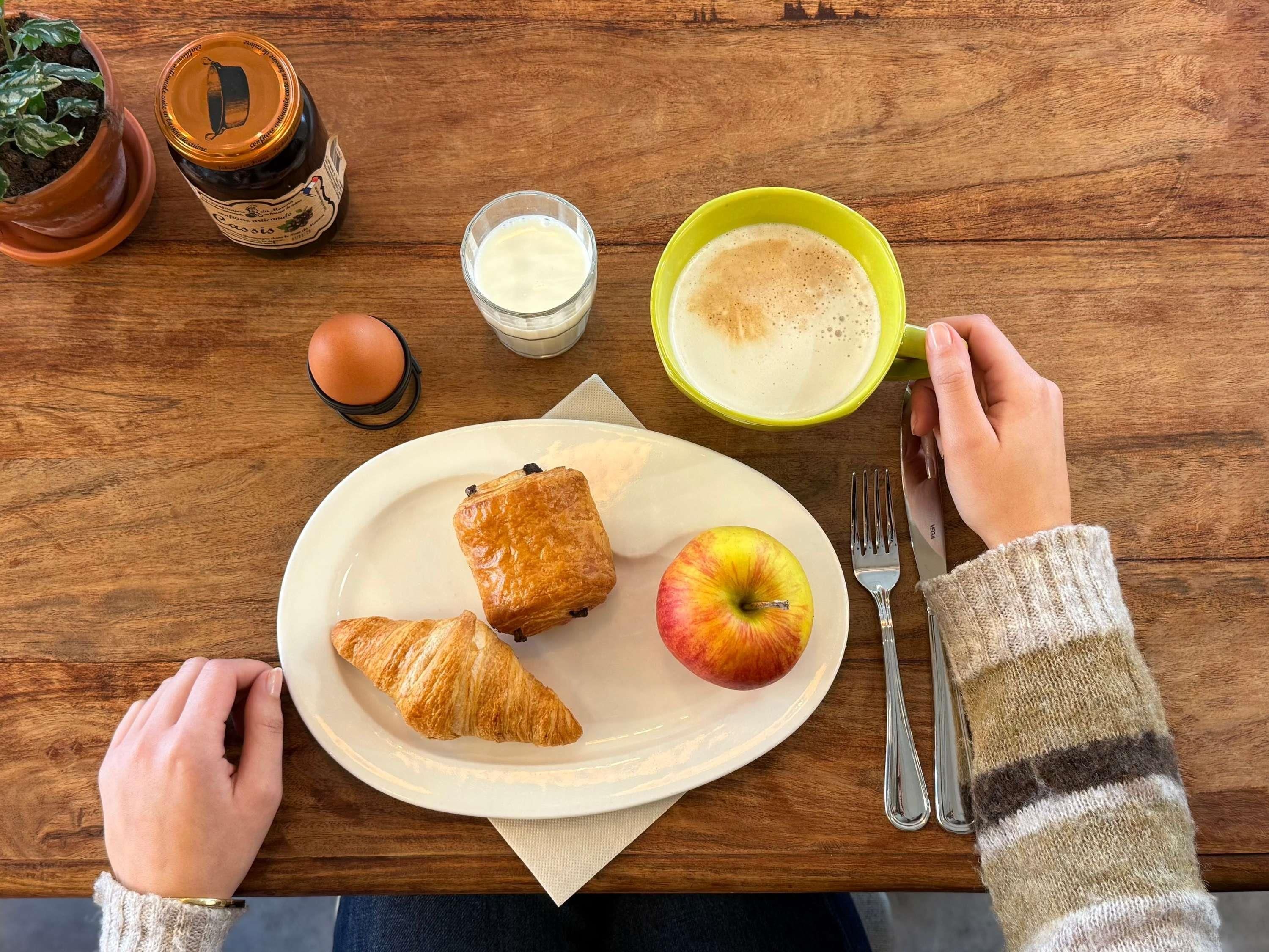 Ein Holztisch mit einem Teller Essen, darunter Croissants und ein Apfel, ein Glas Milch und eine Tasse Kaffee.