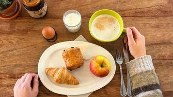Ein Holztisch mit einem Teller Essen, darunter Croissants und ein Apfel, ein Glas Milch und eine Tasse Kaffee.