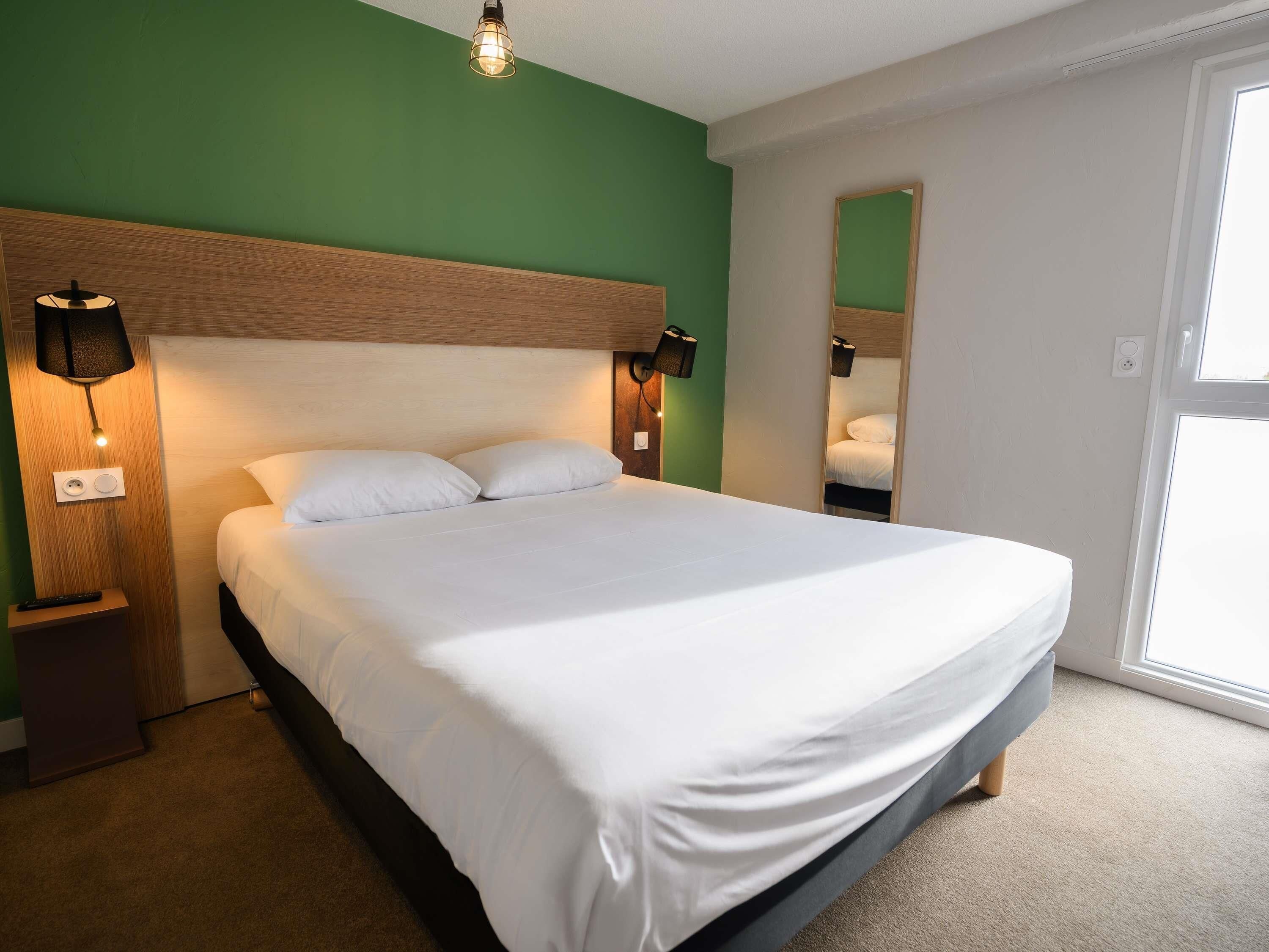 Una cama bien tendida en una habitación con pared verde, cabecera de madera y lámpara de noche.