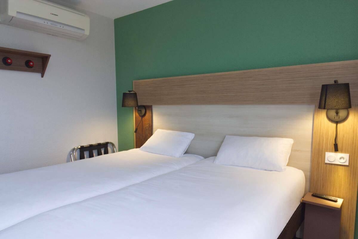 A double bed with white linens, two bedside lamps, a wooden headboard, a wall-mounted air conditioner, and a small wooden nightstand with a remote control.