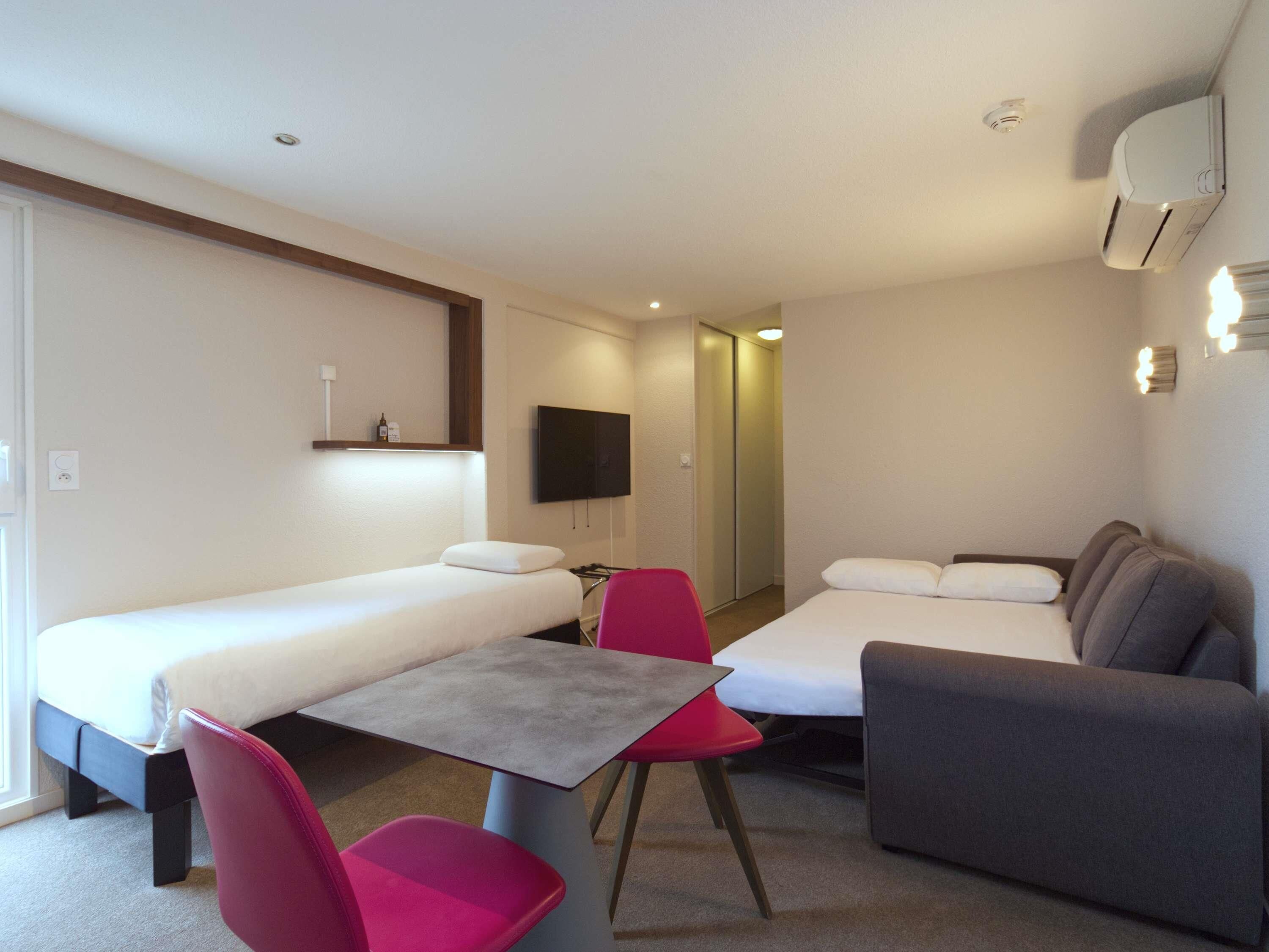 Ein kompaktes Hotelzimmer mit einem Einzelbett, einem kleinen Tisch, zwei Stühlen, einer Couch, einem Fernseher und einer Klimaanlage.
