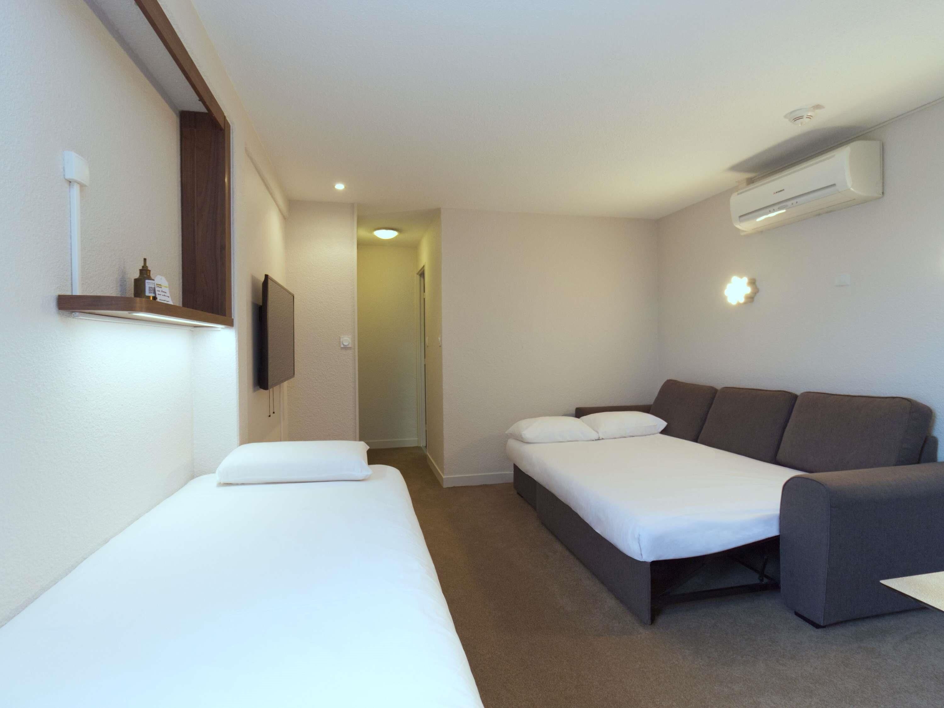 Habitación de hotel con dos camas individuales, un sofá, un equipo de aire acondicionado y un pequeño estante con artículos de aseo.
