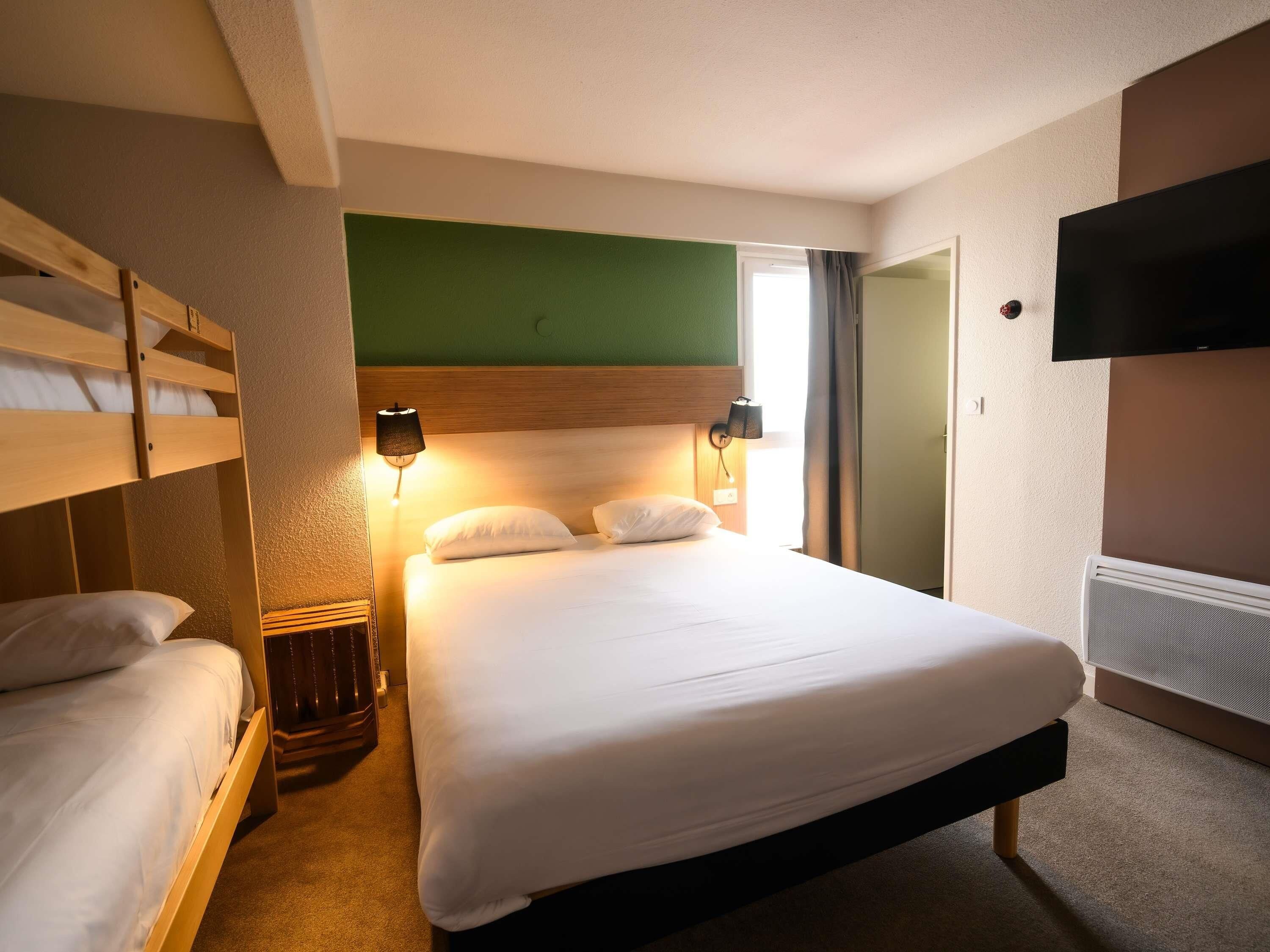 Ein Hotelzimmer mit Etagenbett, einem Einzelbett, einem wandmontierten Fernseher und einem Nachttisch.