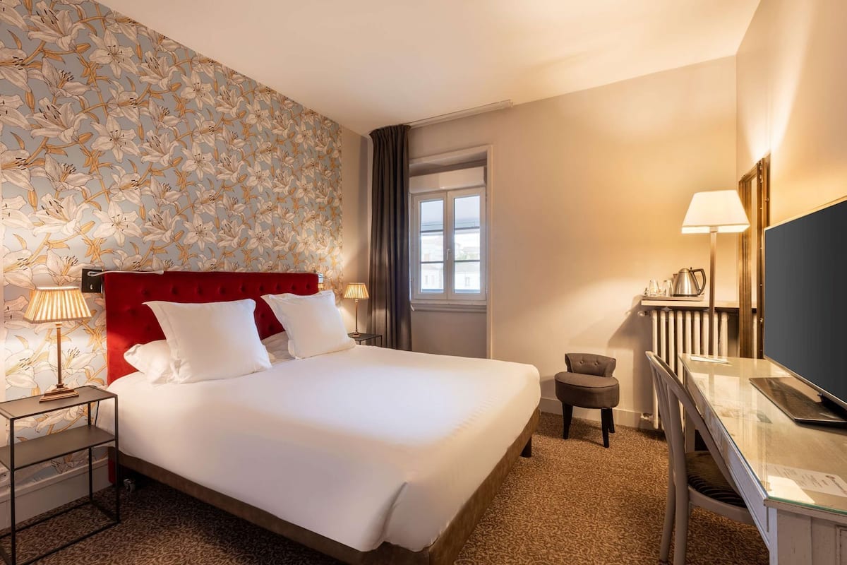 A hotel room with a large bed, a desk with a TV, a chair, and a window.