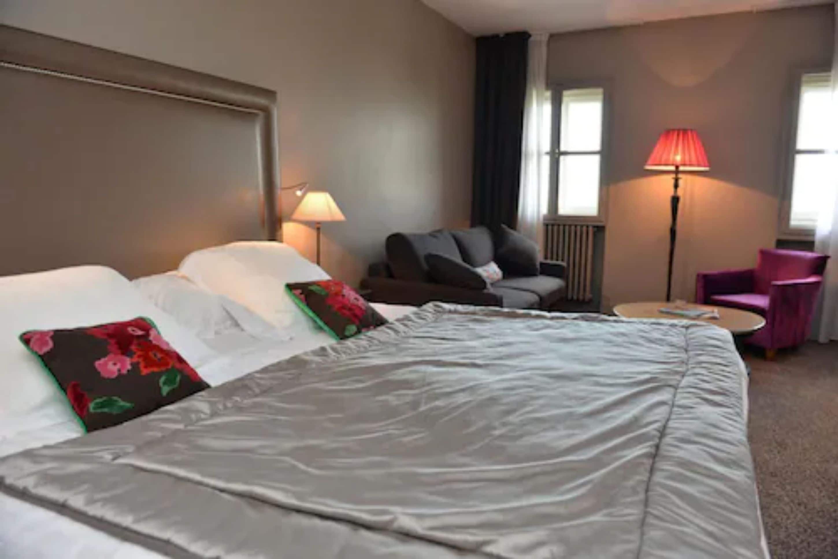 A hotel room with a large bed, a chair, a small table, and a lamp.