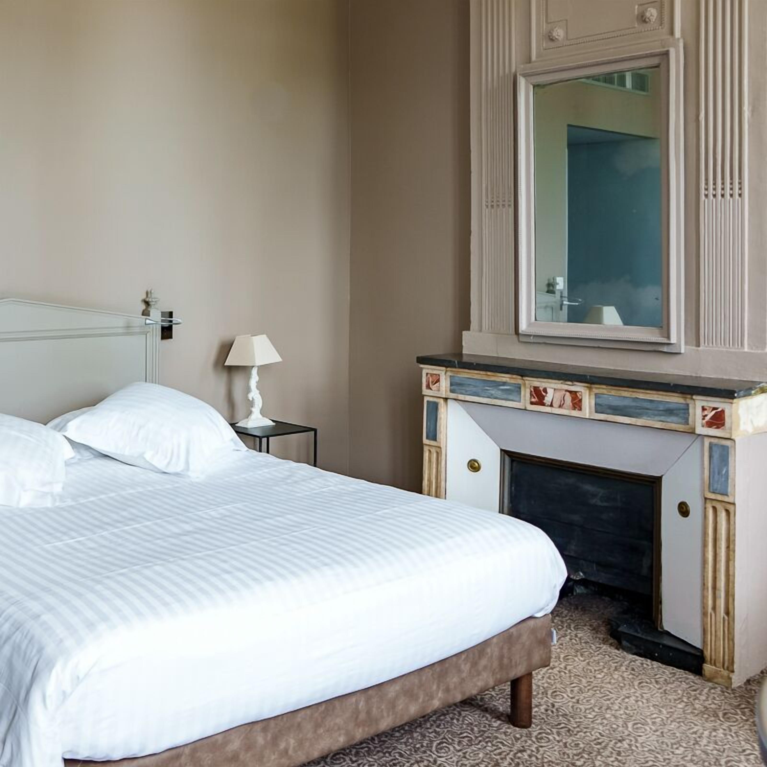 A hotel room with a bed, a nightstand with a lamp, a fireplace, and a mirror above it.