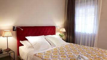 Una habitación de hotel con un cabecero tapizado rojo, ropa de cama blanca y una colcha estampada. Hay una mesita de noche con una lámpara y un reloj.