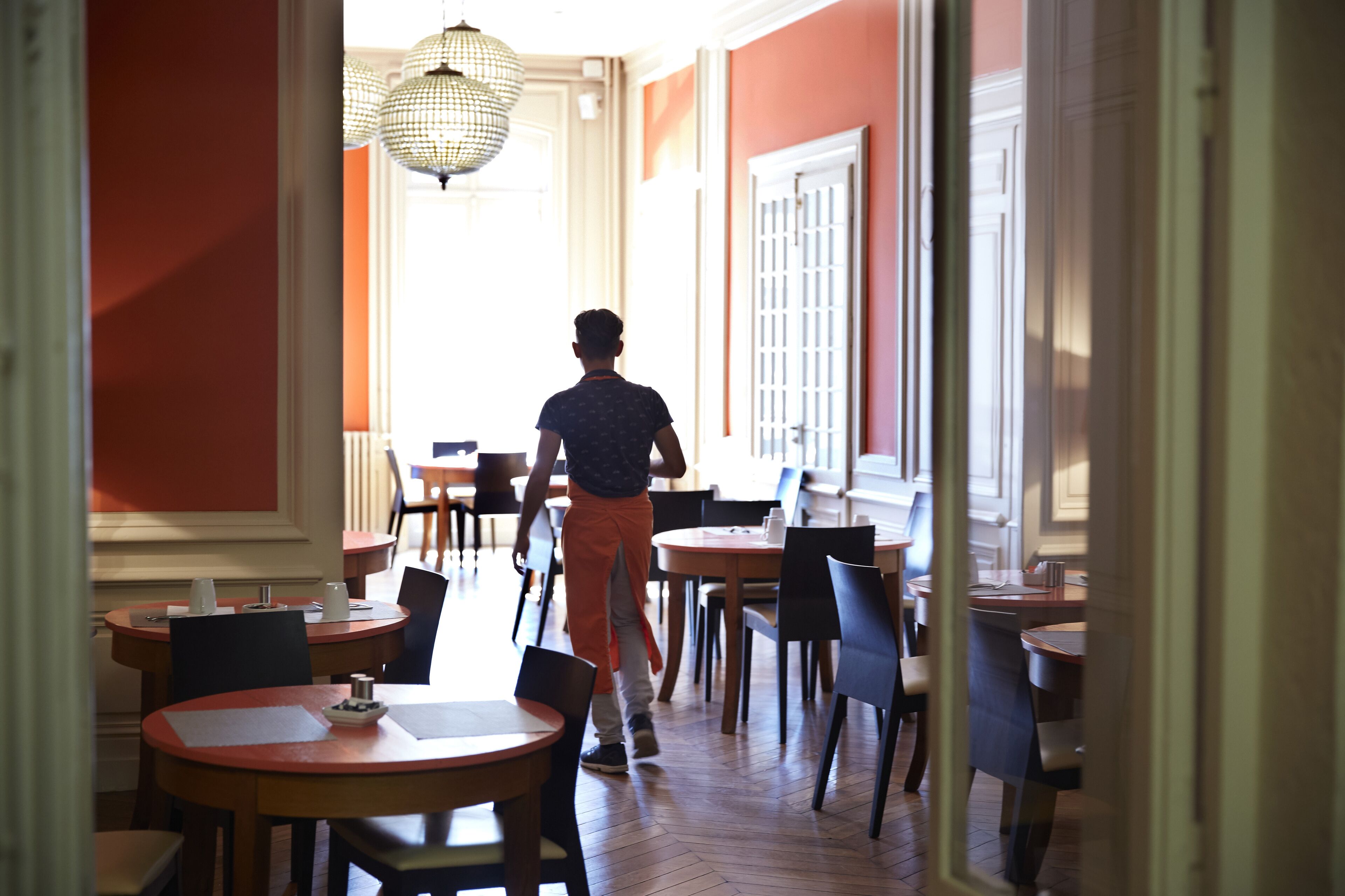 A person walking through a dining area with multiple tables and chairs, a chandelier, and a view of the outside.