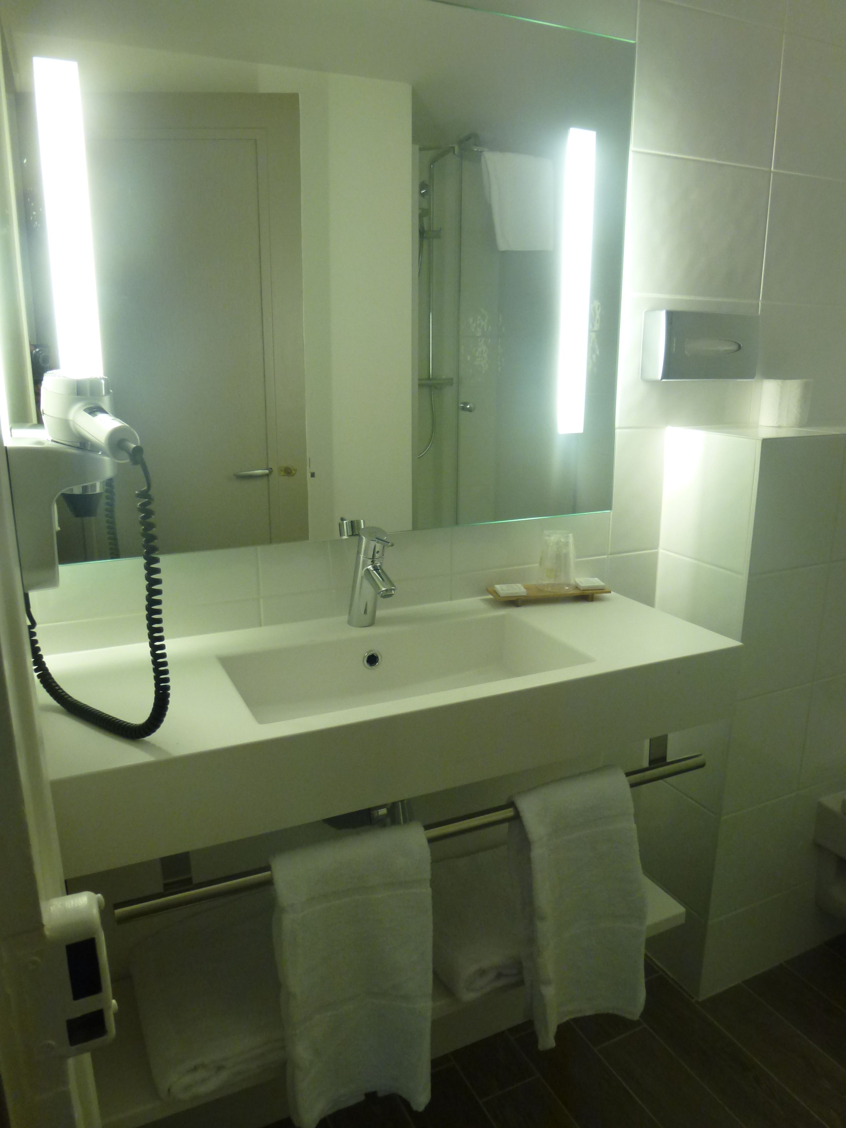 Un baño con un lavabo blanco, un espejo, un secador de pelo de mano y dos toallas colgadas en un perchero.