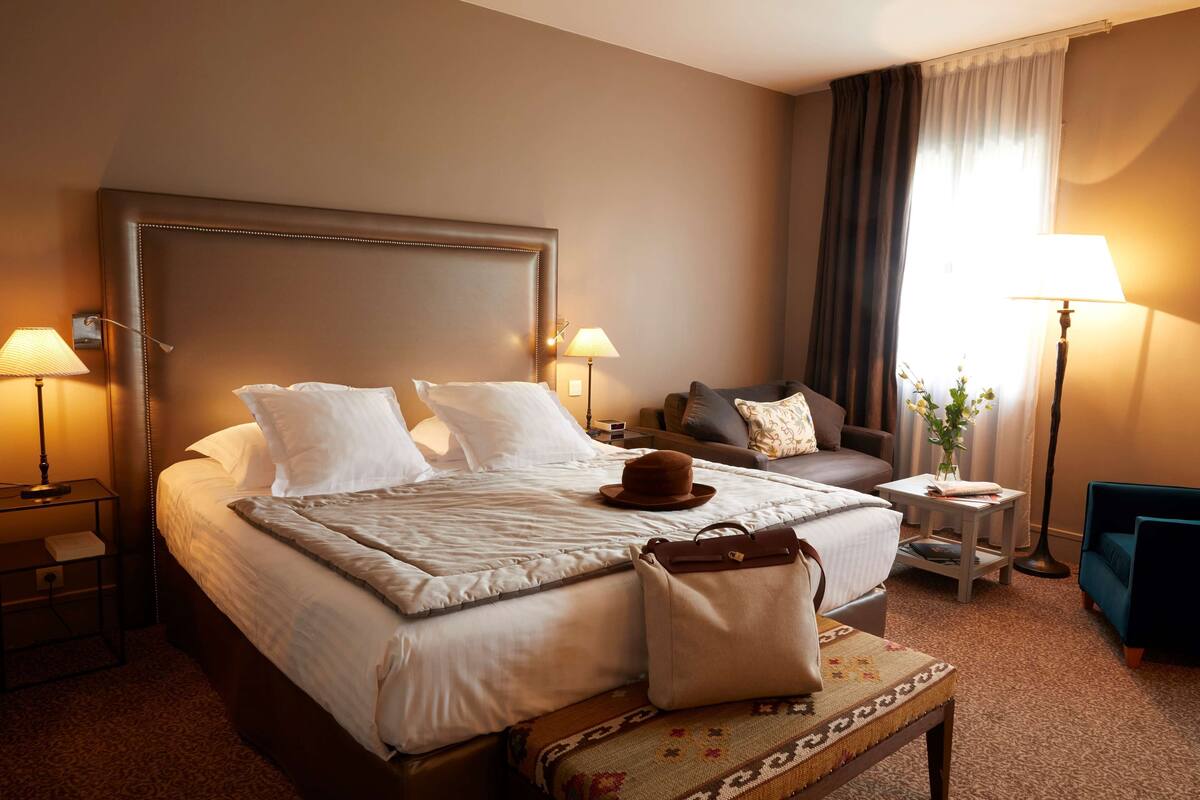 A hotel room with a large bed, two bedside tables, a nightstand, a sofa, a small table with a vase, and a chair.