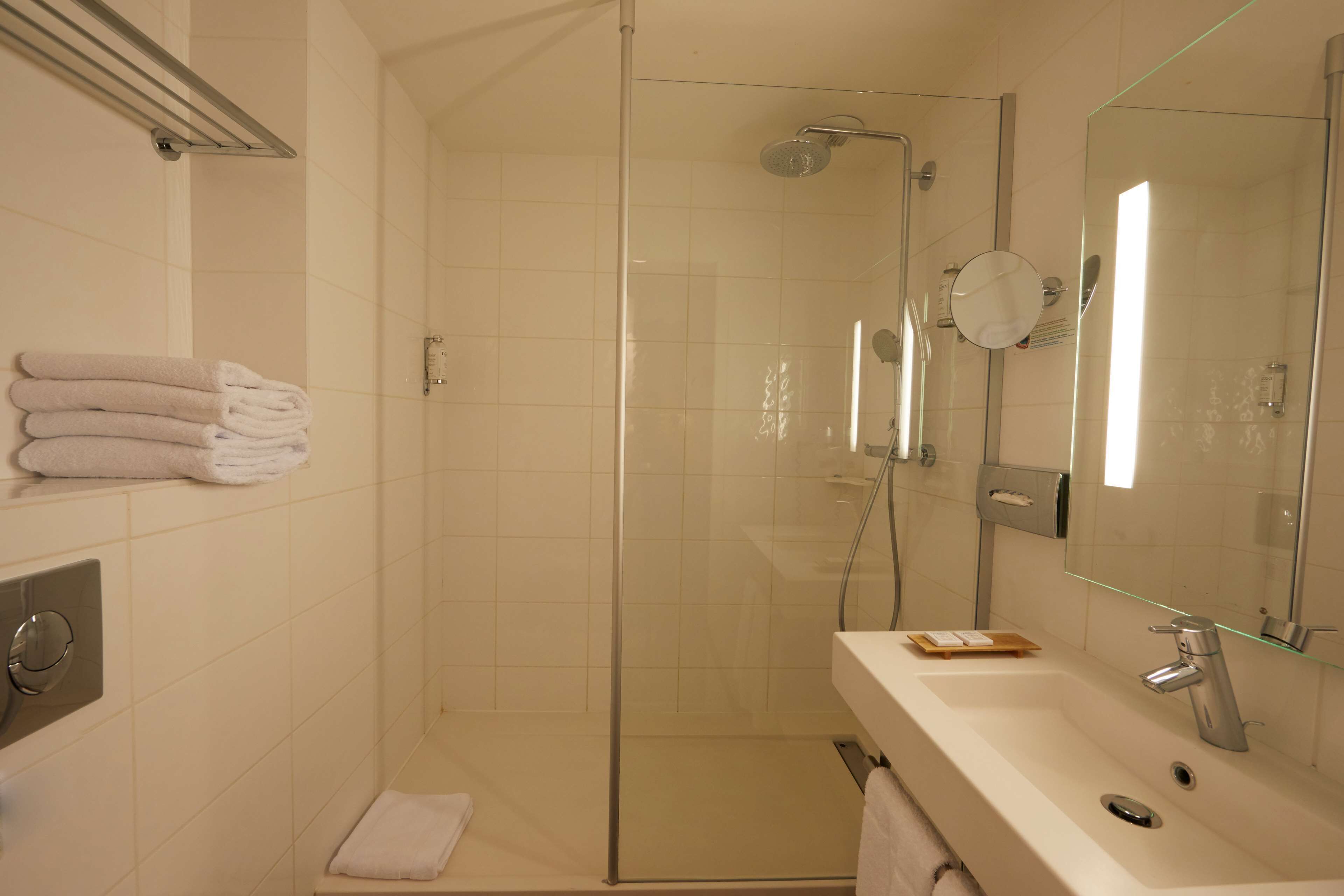 Un baño moderno con ducha envuelta en vidrio, lavabo con espejo y toallas ordenadas en un estante.