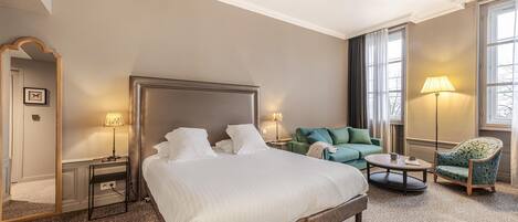 A hotel room with a large bed, a green sofa, a patterned armchair, a small round table, and two bedside lamps.