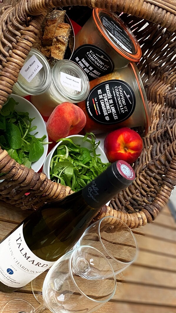 Una cesta con una botella de vino, copas y diversos alimentos, entre ellos frutas y frascos.