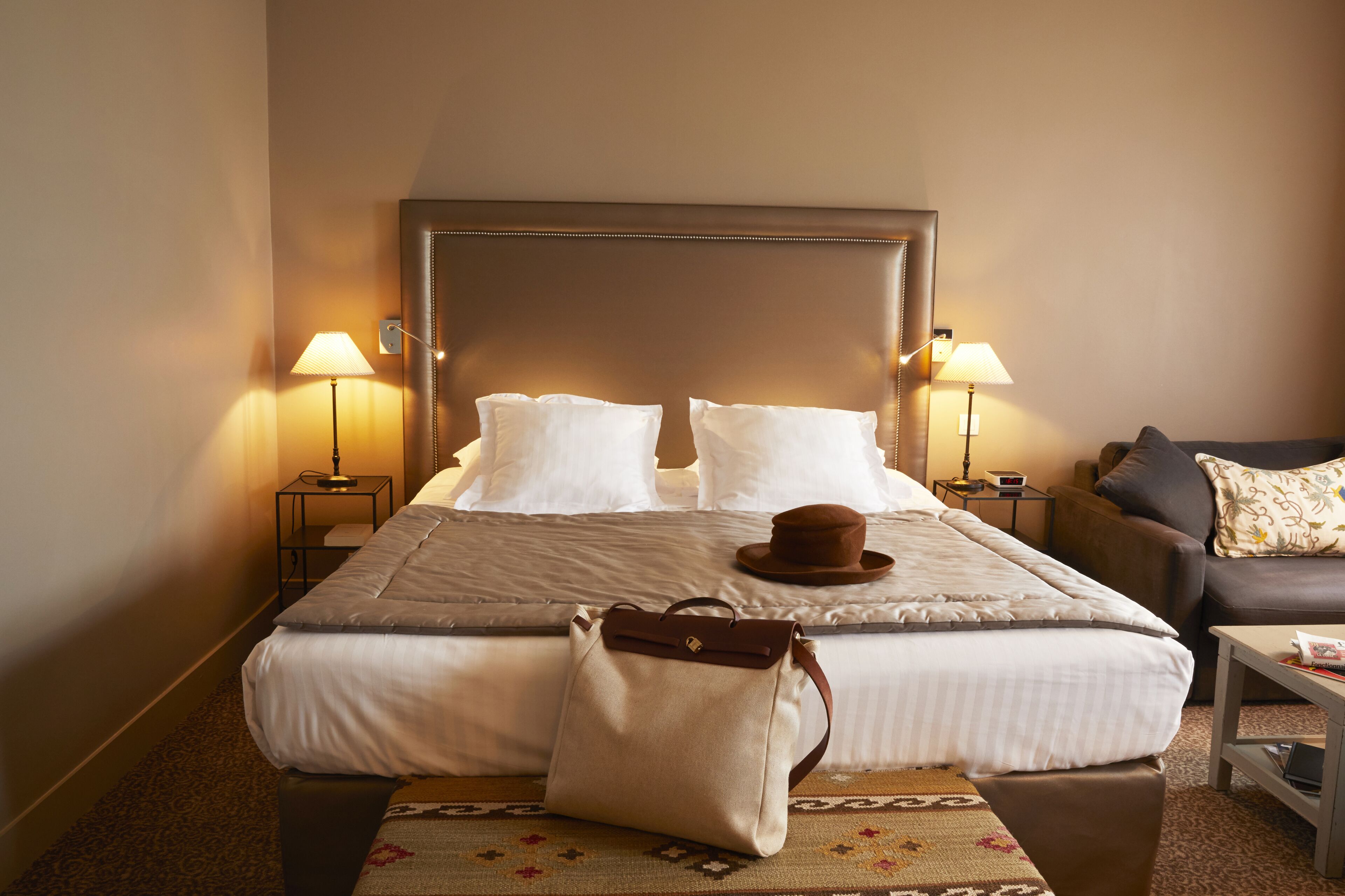 A hotel room with a large bed, two bedside tables with lamps, a brown leather sofa, and a patterned rug.