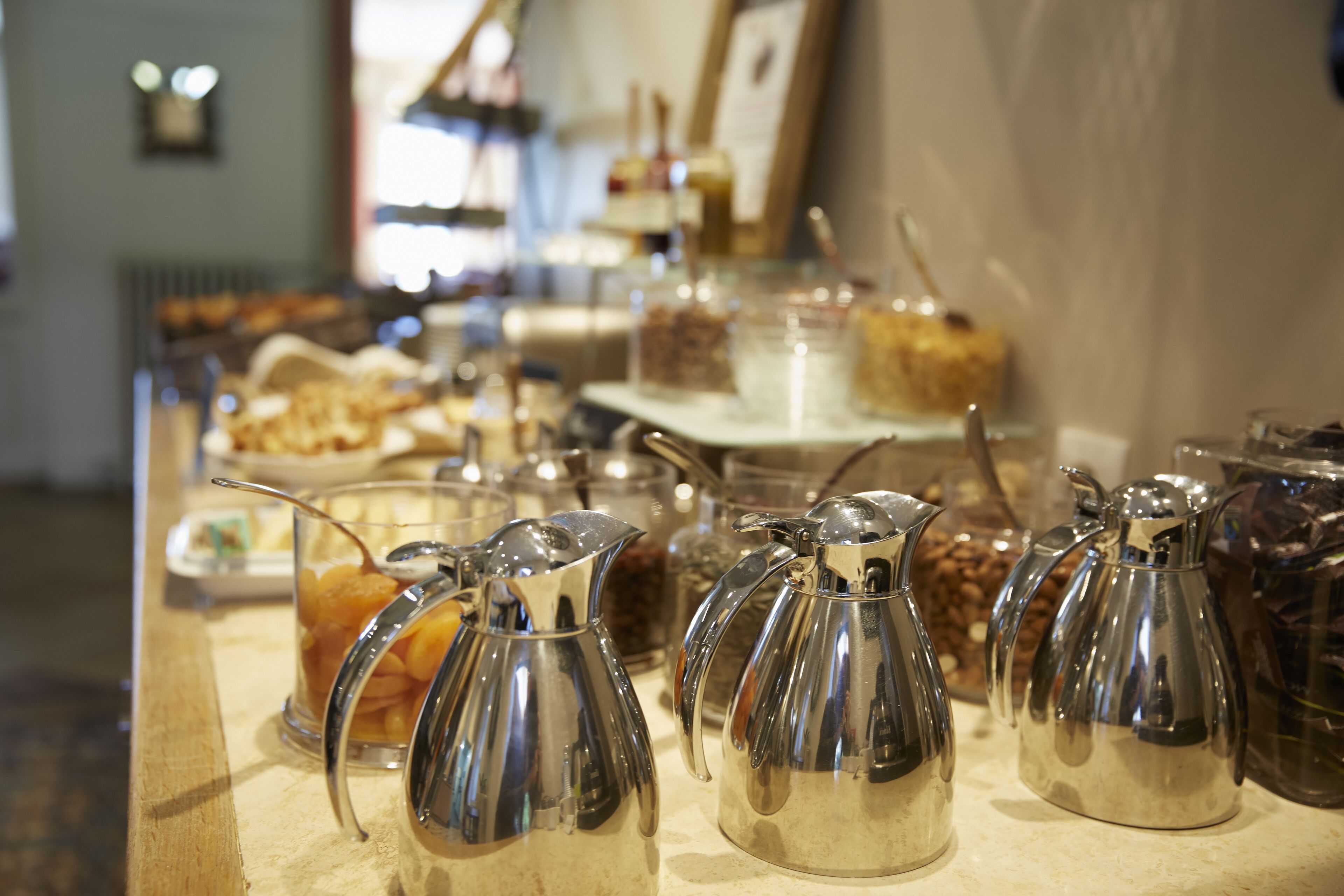 A kitchen counter with multiple stainless steel coffee pots and various food items in the background.