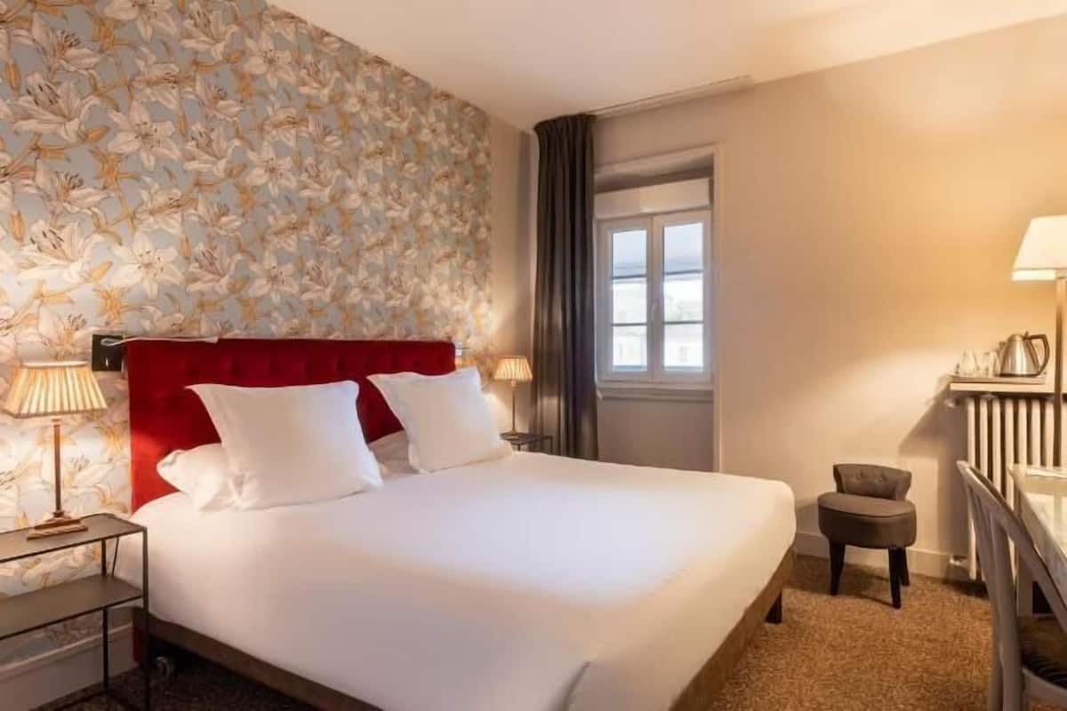 A hotel room with a bed, a desk with a lamp, a chair, a TV, and a window.