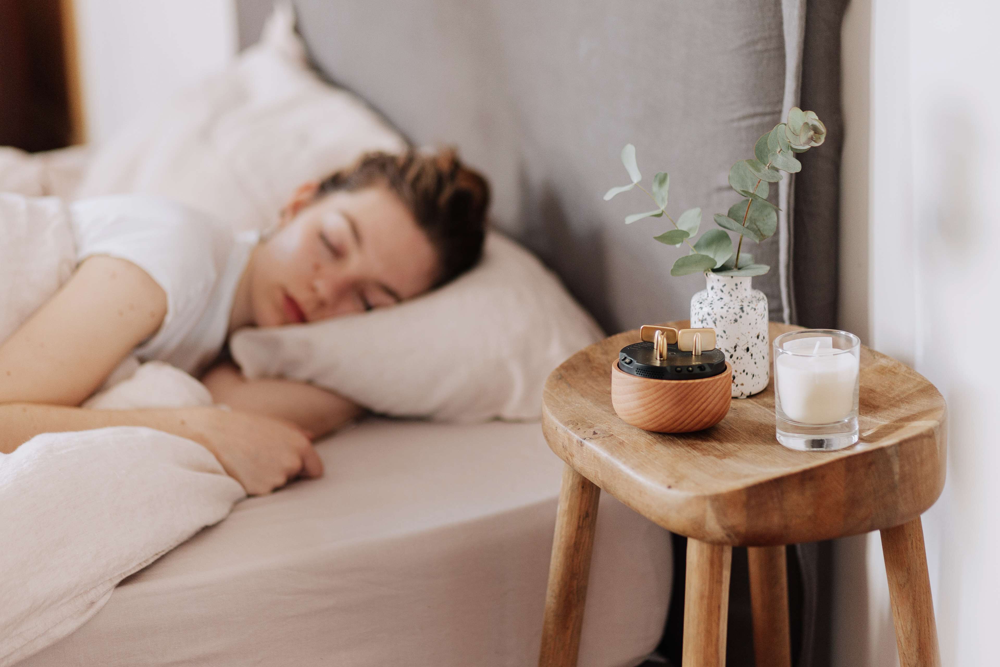 Une personne dort dans un lit, à côté duquel se trouve une table de chevet en bois, sur laquelle sont posés une bougie et une petite plante.