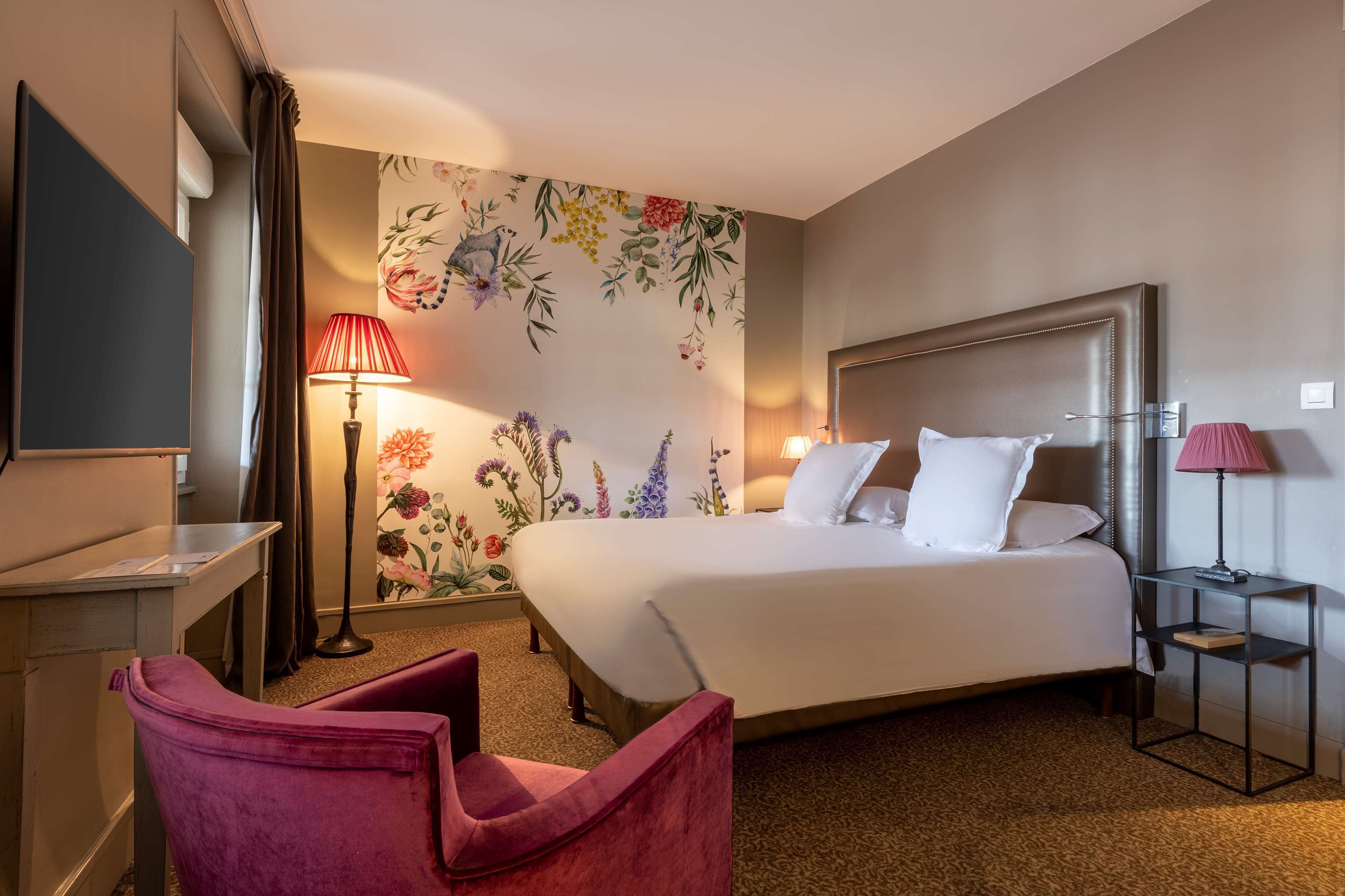 A hotel room with a large bed, a desk, a chair, a TV, and a wall with floral wallpaper.