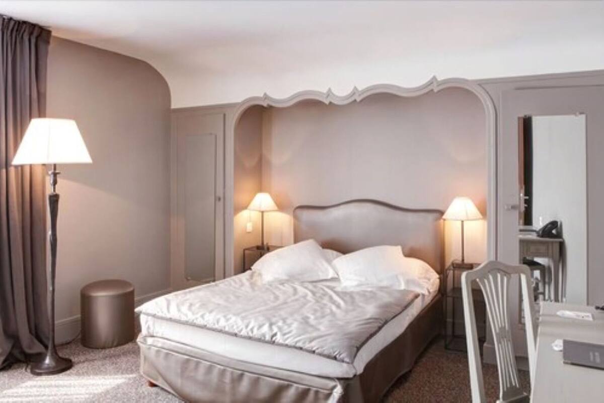 A hotel room with a bed, two bedside lamps, a round bedside table, a mirror, and a door.