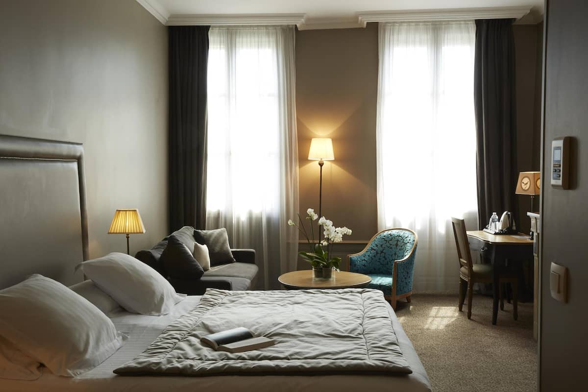 A hotel room with a bed, a desk, a chair, a lamp, and a window with curtains.
