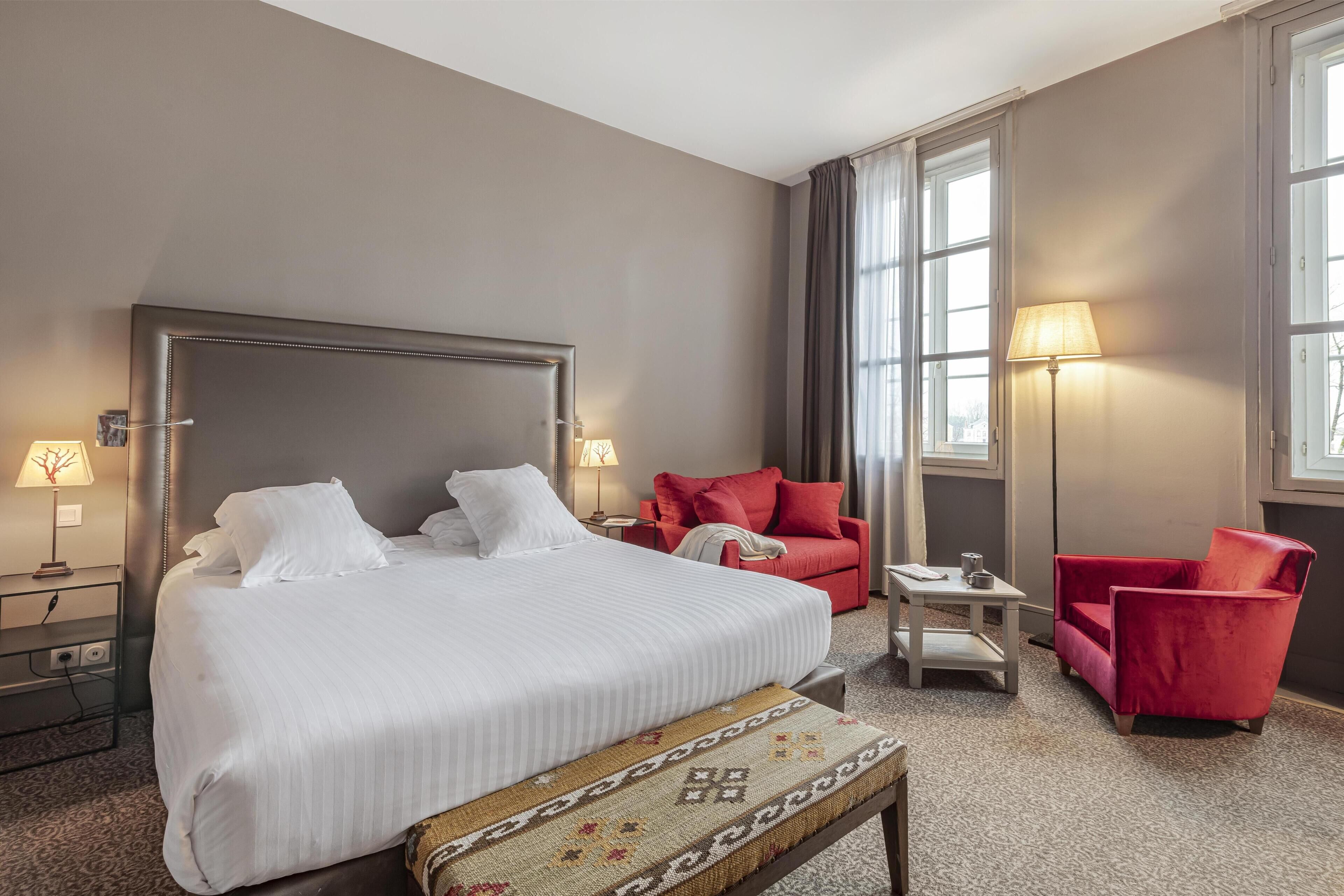 Una habitación de hotel con una cama grande, dos sillones rojos, una mesita y una lámpara.
