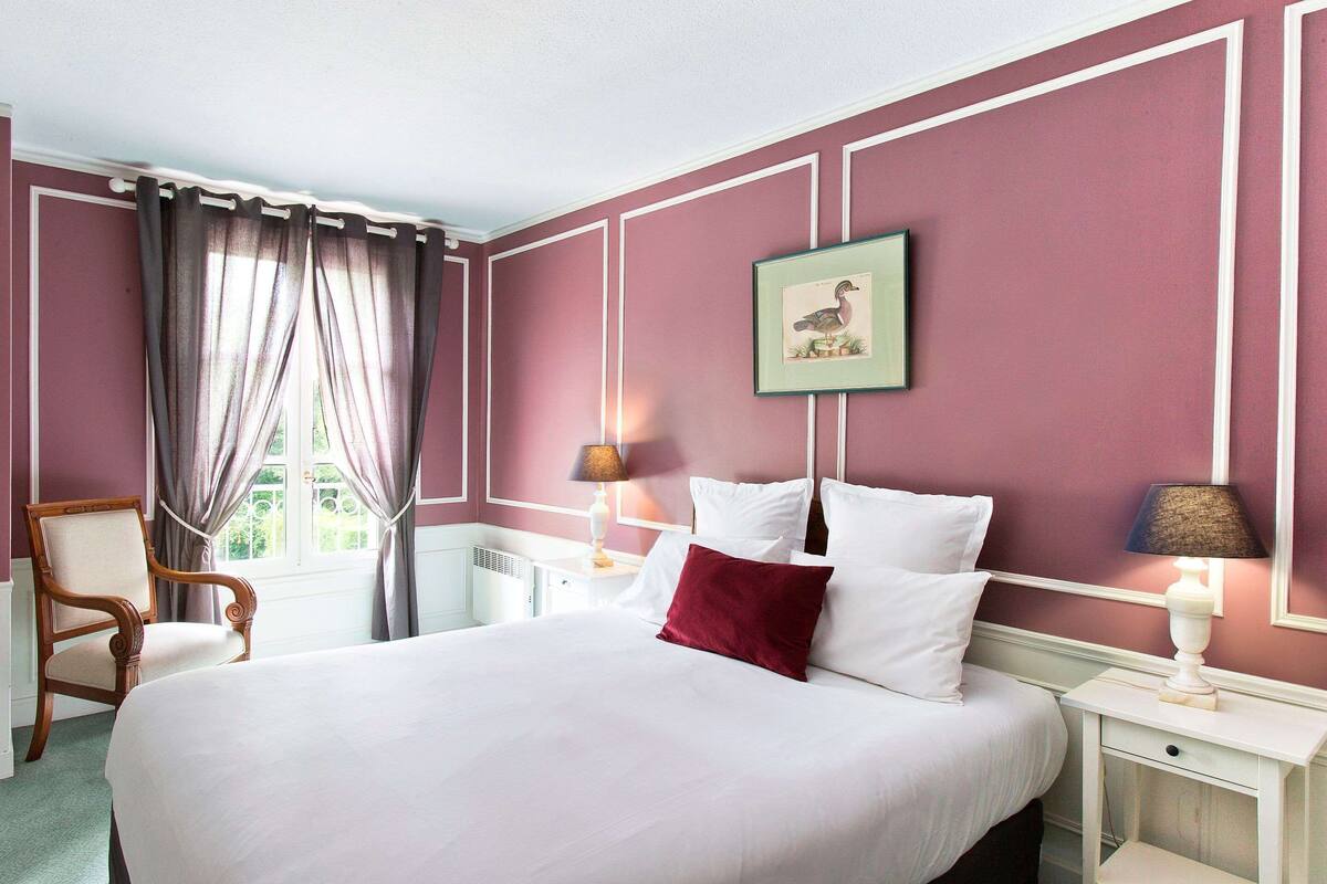 A bedroom with a large bed, a chair, a bedside table with a lamp, and a window with curtains.