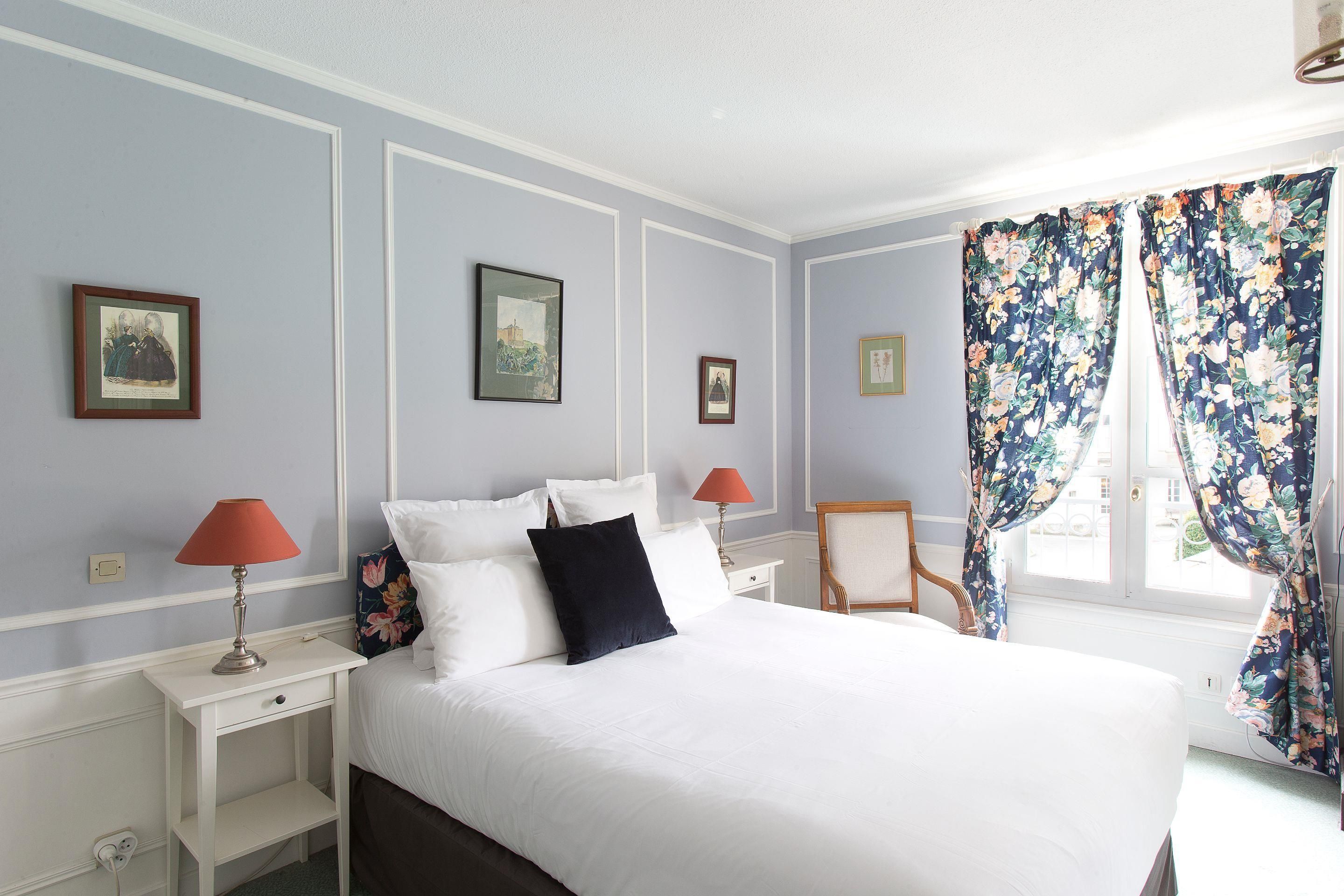 Een slaapkamer met een groot bed, twee nachtkastjes, een stoel en een raam met bloemen gordijnen.