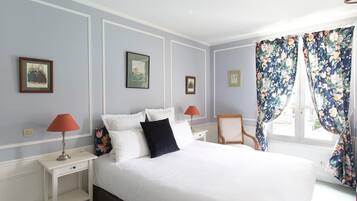 Een slaapkamer met een groot bed, twee nachtkastjes, een stoel en een raam met bloemen gordijnen.
