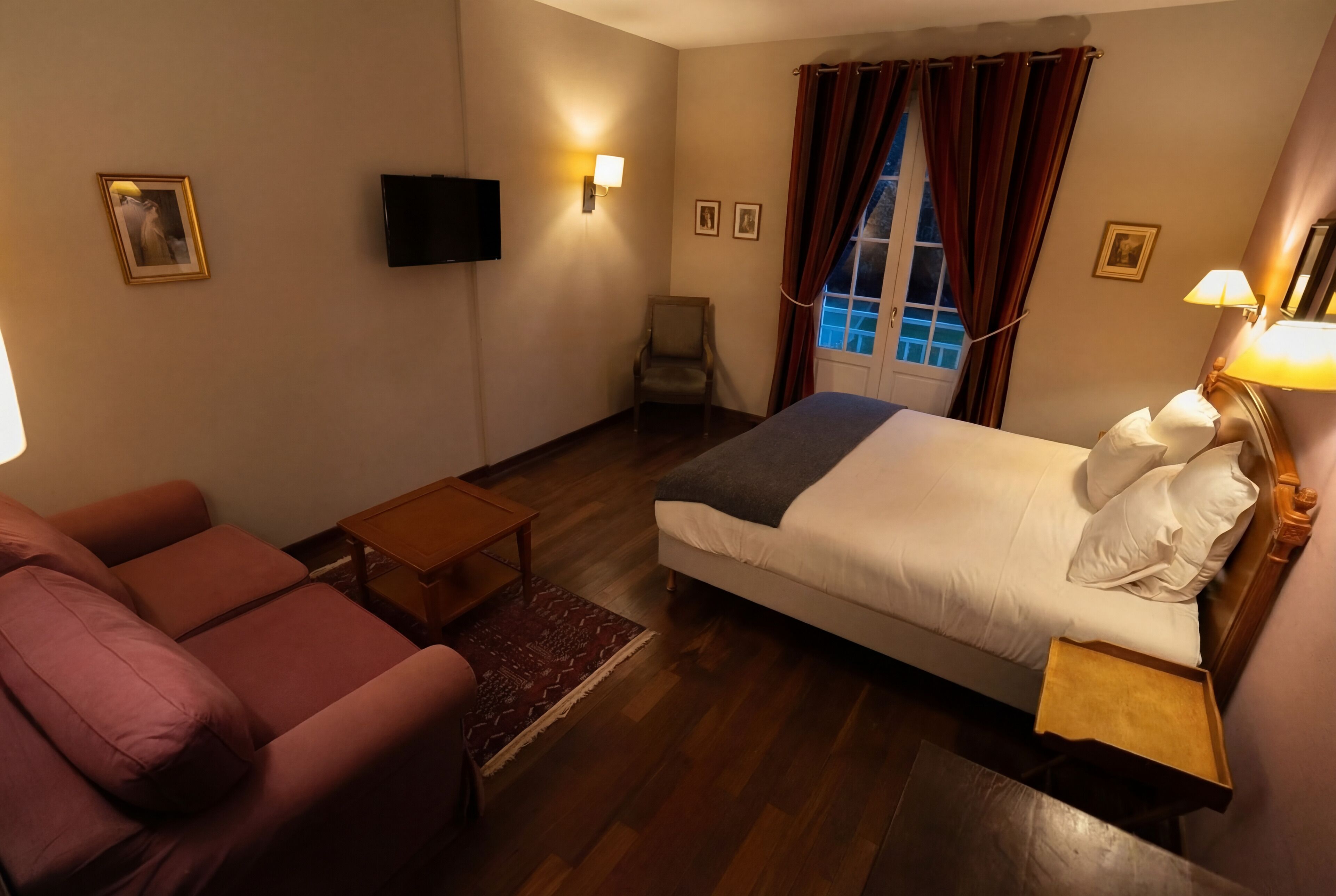A hotel room with a bed, a sofa, a chair, a small table, and a TV mounted on the wall.