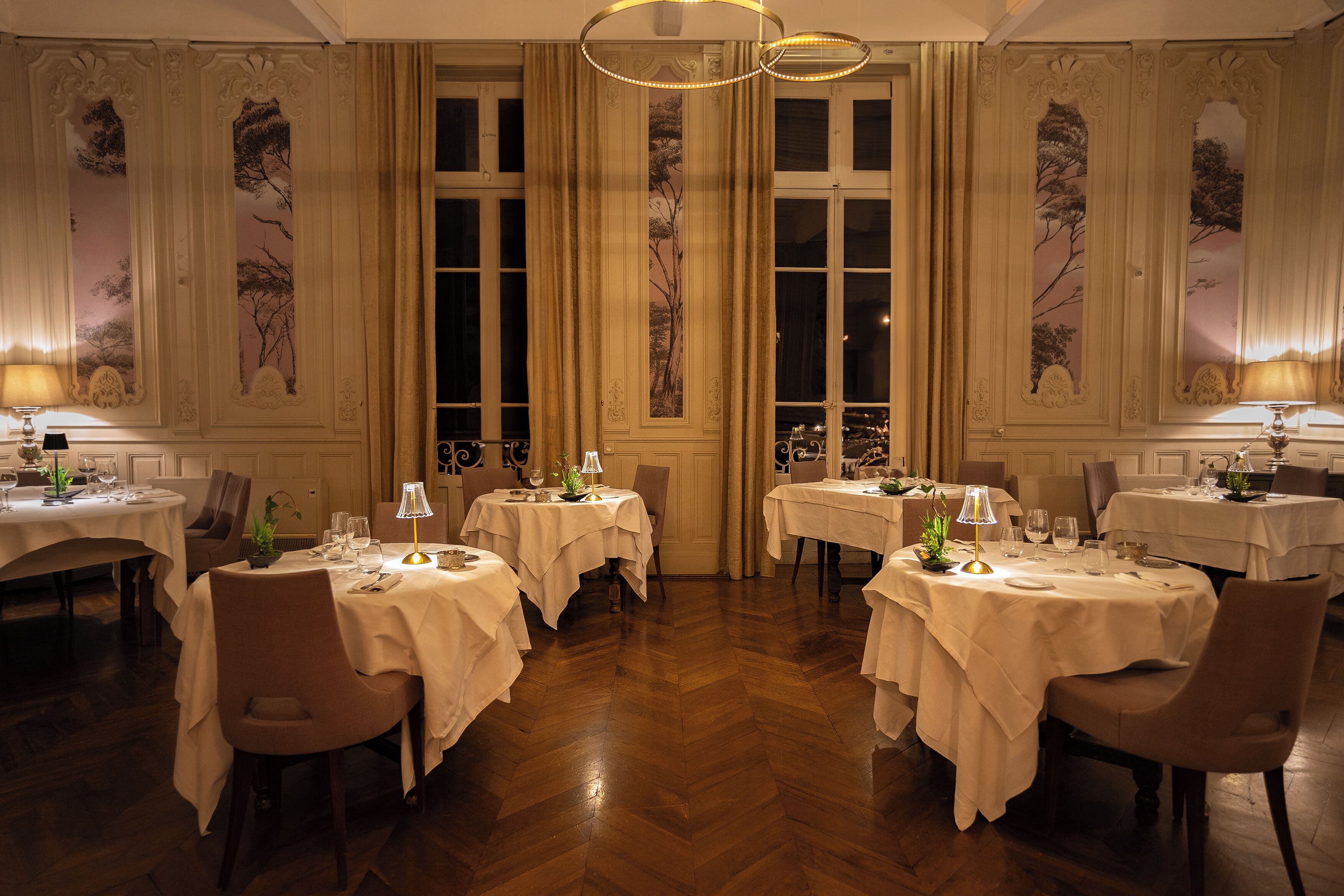 A formal dining room with multiple tables, each set with white tablecloths, chairs, and table lamps.