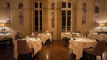 A formal dining room with multiple tables, each set with white tablecloths, chairs, and table lamps.