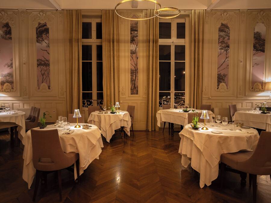 A formal dining room with multiple tables, each set with white tablecloths, chairs, and table lamps.