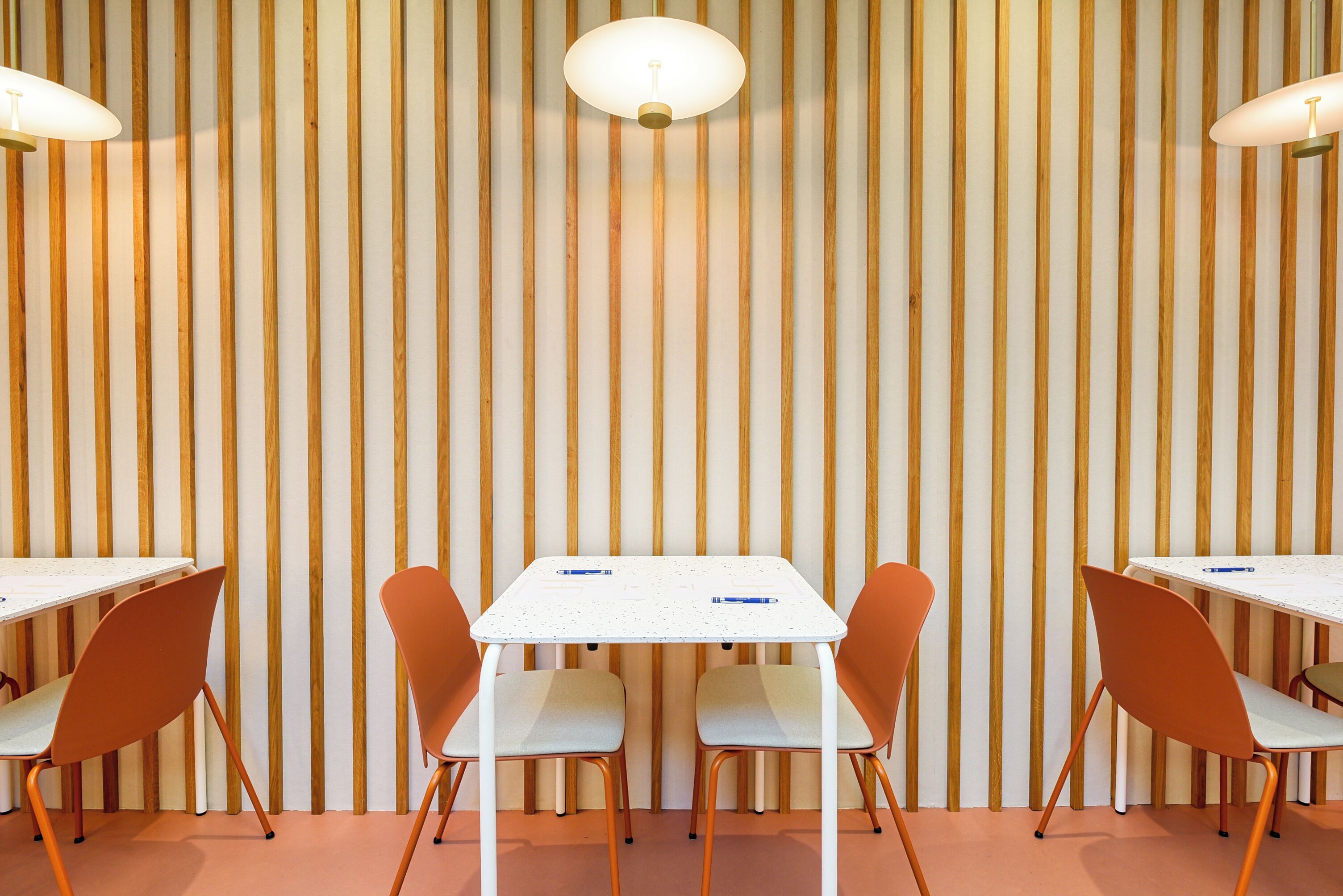 Una habitación con pared de madera a rayas, mesas blancas y sillas naranjas.