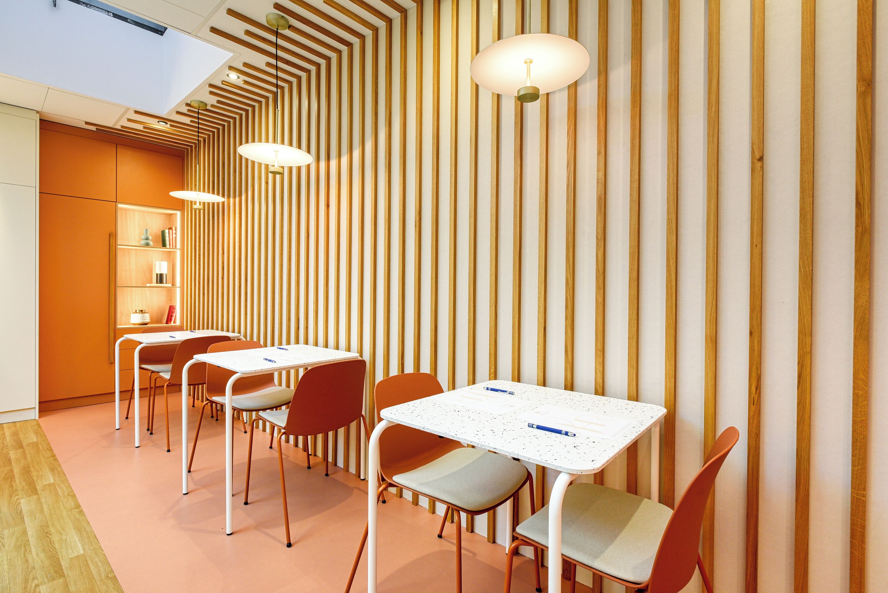 Un comedor moderno con paredes de madera a rayas, mesas blancas y sillas anaranjadas.