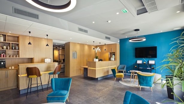 Un moderno vestíbulo de hotel con mostradores de recepción de madera, sillas azules y amarillas, un televisor de pantalla plana y una planta en maceta.