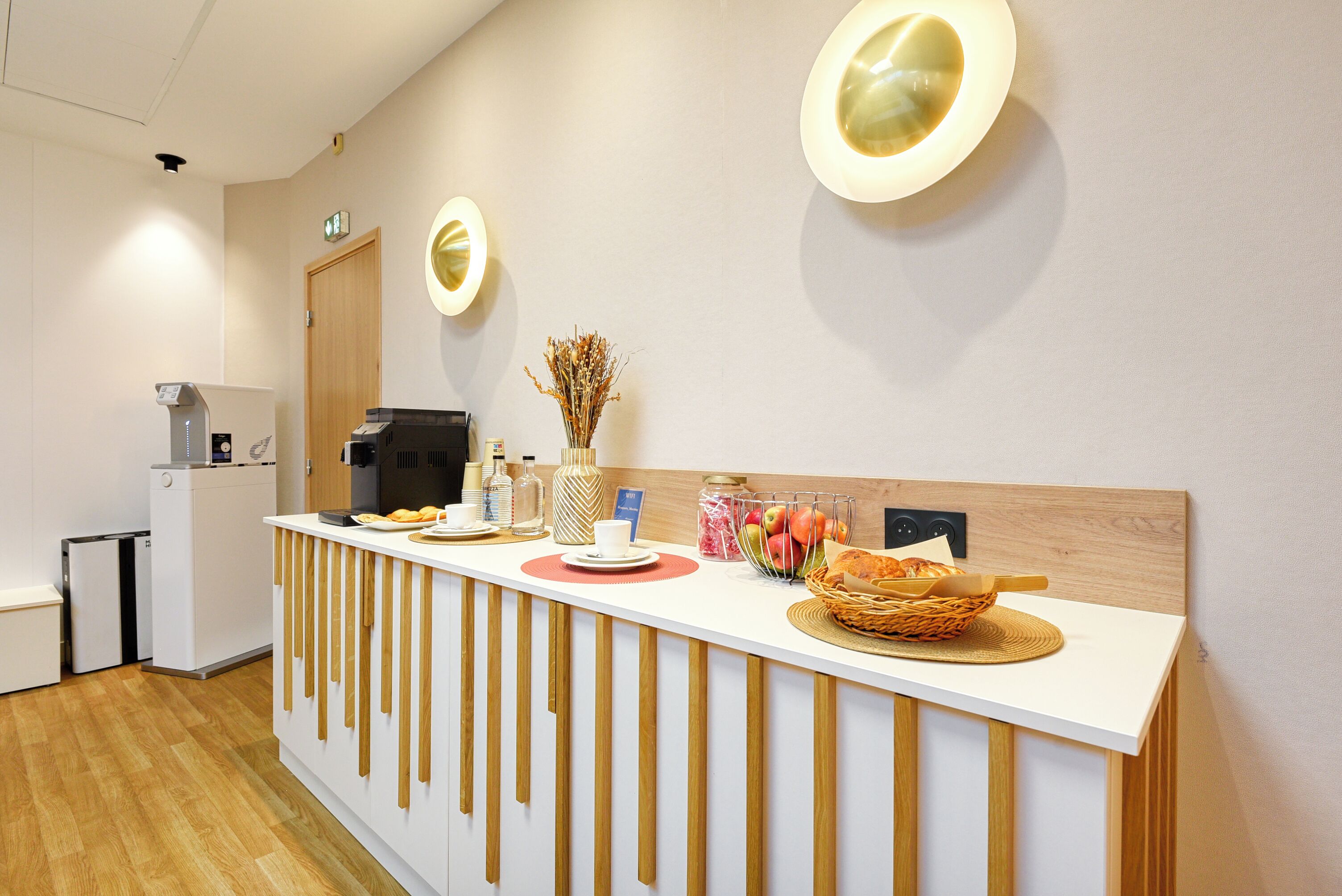 A reception desk with a white countertop, wooden slats, and a basket of bread. There are wall-mounted lights and a door in the background.
