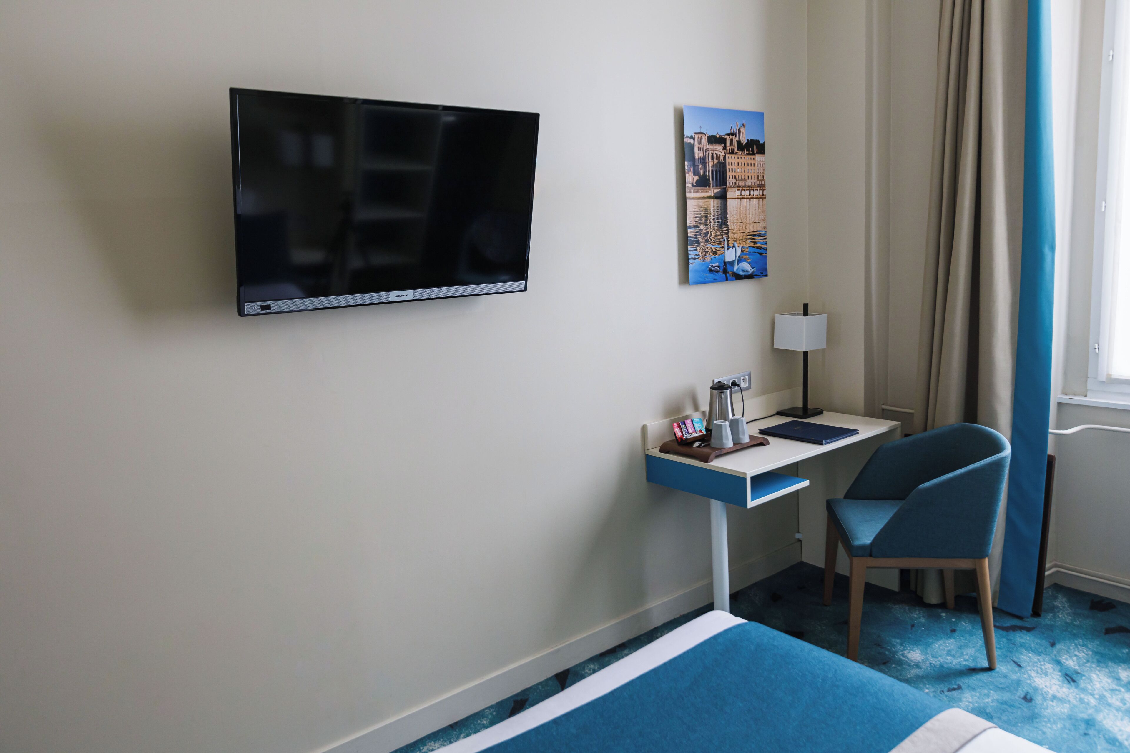 Une chambre d’hôtel dotée d’un téléviseur à écran plat fixé au mur, d’un bureau avec une lampe, d’une chaise et d’un lit recouvert d’une couvre-lit à rayures bleues et blanches.