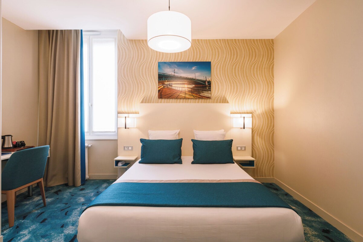 Habitación de hotel con cama, dos lámparas de noche, un escritorio con silla, una ventana con cortinas y un cuadro colgado en la pared.