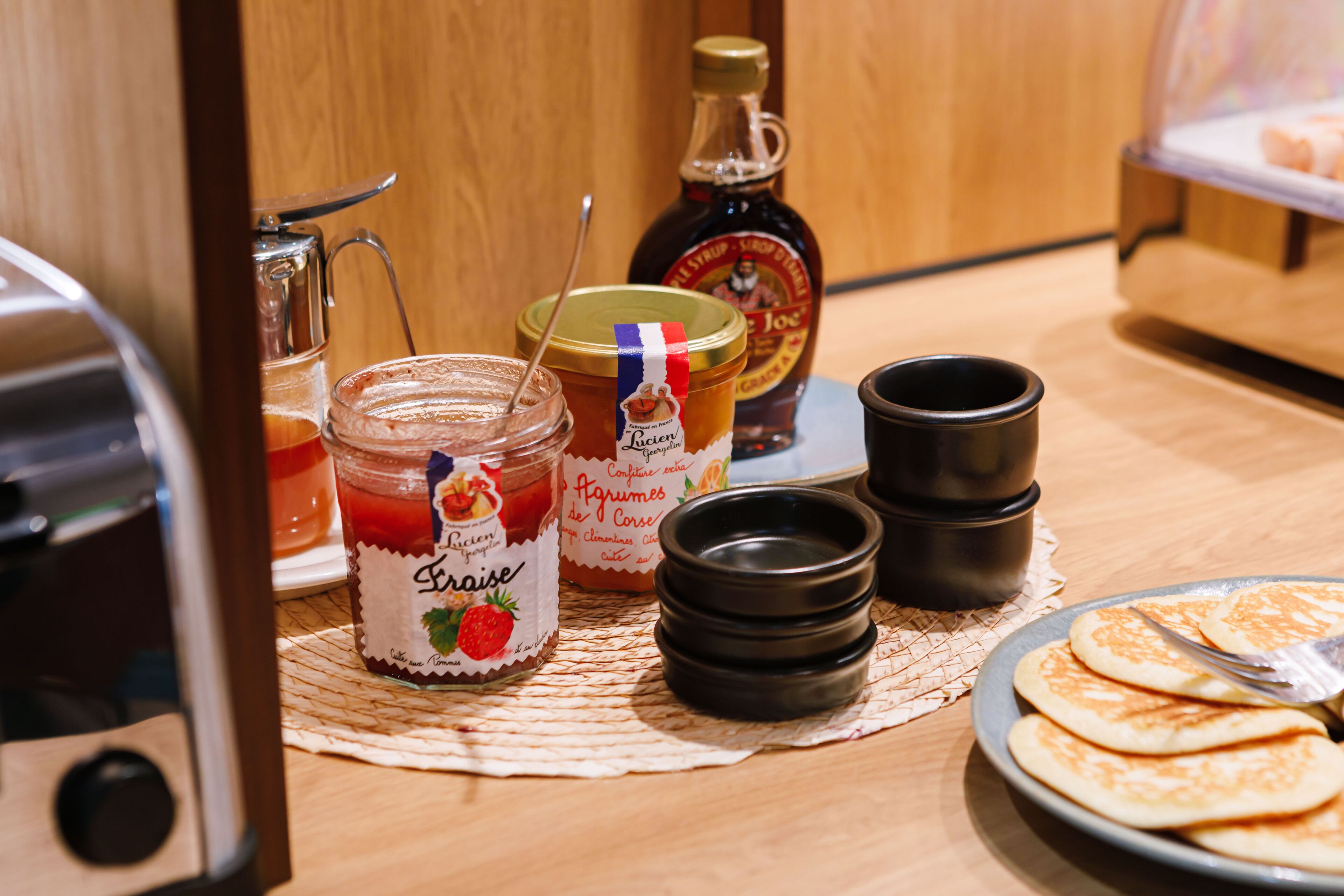 Un comptoir de cuisine avec une assiette de crêpes, un pot de confiture de fraises, une bouteille de sirop et une machine à café.
