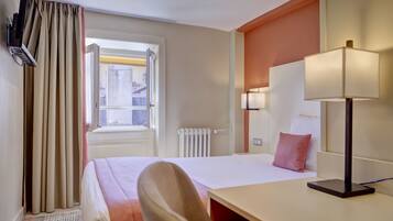 Una habitación de hotel con cama, escritorio, silla, lámpara, ventana con cortinas y radiador.