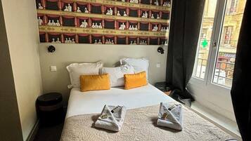 Habitación de hotel con una cama grande, una pared con cuadros enmarcados, una ventana con cortinas y una pequeña mesa redonda.