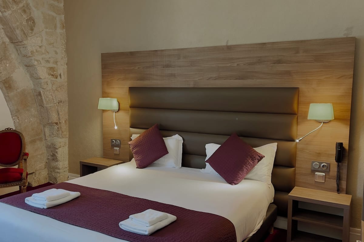 A hotel room with a large bed, two bedside tables, a red chair, and a stone wall.