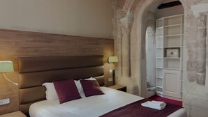 Een hotelkamer met een houten hoofdbord, een bed met wit en paars beddengoed, een bedlamp, een stopcontact en een stenen muur met een inbouwplank.