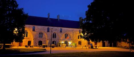 A historic stone building with arched windows and a central entrance, surrounded by a well-maintained lawn and trees.