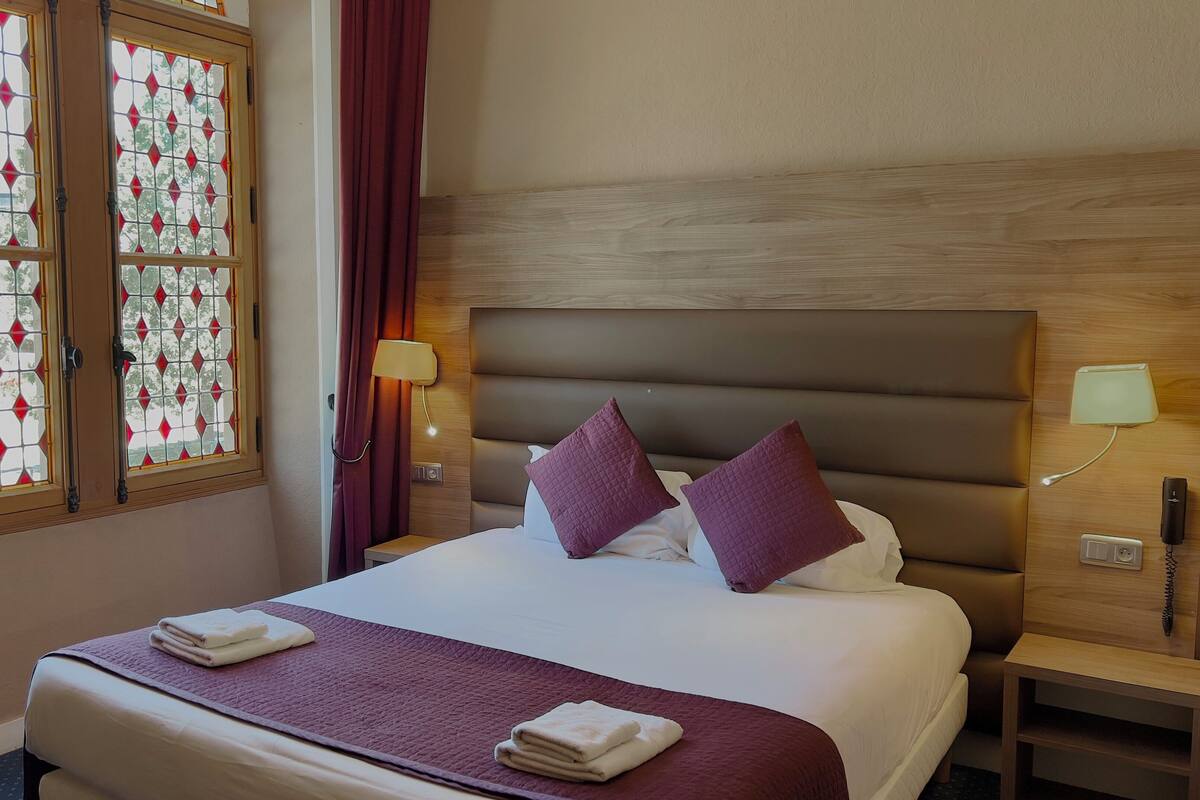 A hotel room with a bed, two bedside lamps, a nightstand, a wooden headboard, and a window with stained glass.